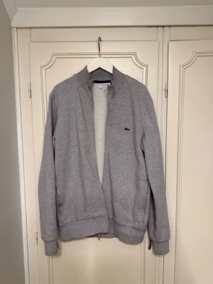 Grå ziptröja från Lacoste - Säljer en grå ziptröja från Lacoste. Perfekt för chill dagar och enkel att matcha med jeans eller joggers. Tröjan är nästan i nyskick!