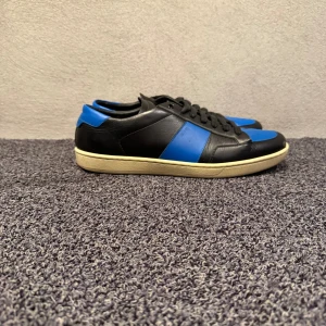 Saint Laurent skor - Säljer ett par snygga sneakers från Saint Laurent med svart och blå ovandel i skinn, vit platt sula och klassisk snörning. Skorna har en rund tå och stilren design med blå detaljer på sidan och hälen. Perfekta för dig som gillar exklusiva märken och streetstyle.