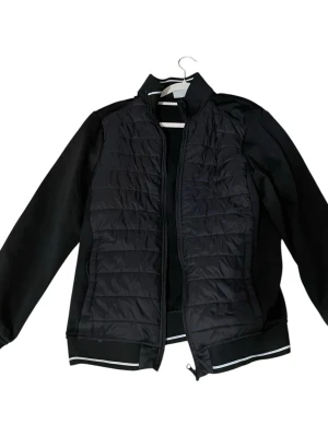 Svart quiltad pufferjacka - Säljer en svart pufferjacka med quiltad front och släta ärmar. Jackan har dragkedja framtill och ribbade muddar med vit rand vid ärmslut och nederkant. Perfekt för dig som vill ha en stilren och sportig look.