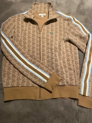 Beige mönstrad ziptröja Lacoste S - Säljer en beige ziptröja från Lacoste med brunt geometriskt mönster och blå/vita ränder längs ärmarna. Tröjan har ståkrage, dragkedja framtill och ribbade muddar. Ikoniska Lacoste-loggan på bröstet. Regular fit, perfekt för chill och streetwear.