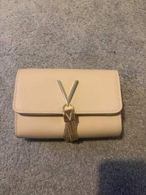 Beige axelväska med guldkedja från Valentino - Snygg beige axelväska från Valentino med guldfärgad kedja och dekorativt V-spänne framtill. Väskan har en stilren, rektangulär form och är tillverkad i skinnimitation. Insidan har fack och dragkedja för småsaker. Perfekt för dig som gillar stilrena detaljer och lyxig känsla.