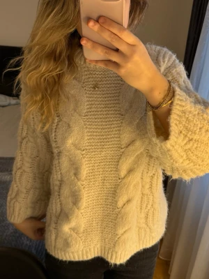 Beige/vit stickad tröja från Vero Moda - Supermysig beige stickad tröja från Vero Moda med grova flätstickade detaljer framtill och på ärmarna. Tröjan har rund hals och långärmad passform, perfekt för kyliga dagar. Materialet är mjukt och fluffigt, vilket gör den extra skön att bära. Ger mig story Gilmore vibes!