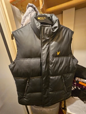 Svart dunväst från Lyle & Scott - Säljer en svart dunväst från Lyle & Scott med gul broderad logga på bröstet. Västen har hög krage, två fickor med dragkedja och quiltad design. Perfekt att slänga över en hoodie när det är kyligt ute.