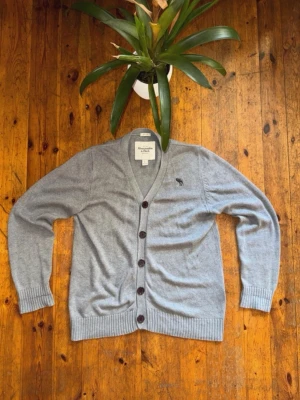 Grå kofta från Abercrombie & Fitch - Snygg grå kofta från Abercrombie & Fitch i storlek L. Koftan är stickad i 100% bomull, har v-ringning och knäpps med fem mörka knappar framtill. Diskret logga broderad på bröstet och ribbade muddar vid ärmslut och nederkant.