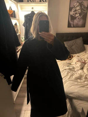 Mörkblå ullkappa med bälte - Säljer en stilren mörkblå kappa i ull med bälte i midjan. Kappan har lång ärm och klassisk krage, perfekt för dig som gillar en clean look. Materialet är mjukt och värmande, och färgen är djup mörkblå. Passformen är normal och kappan går ner till knäna.