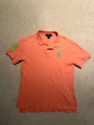 Orange piké från Ralph Lauren - Säljer en orange kortärmad polo från Ralph Lauren med klassisk krage och två knappar. Stor grön logga broderad på bröstet och en grön siffra på ärmen. Pikén har ett litet håll vid taggen, men göms lite gran bakom kragen från utsidan. Hör av er vid intresse! (Pris går att diskutera)🤝