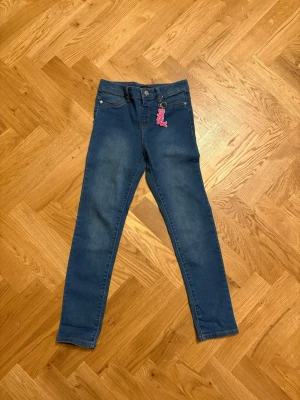 DKNY Blå skinny jeans strl 10 - Snygga blå skinny jeans med klassisk femficksdesign och en cool rosa nyckelring som detalj vid midjan. Jeansen har smal passform och är tillverkade i stretchigt jeansmaterial för extra komfort. Perfekta för dig som gillar en enkel och stilren look.