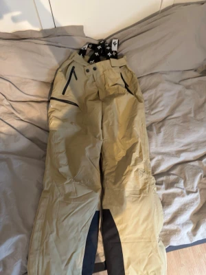 Beige skidbyxor från Cross Sportswear - Säljer ett par beige skidbyxor från Cross Sportswear i storlek L. Byxorna har svarta förstärkningar vid bensluten, svarta dragkedjor på fickorna och justerbara hängslen med logotyp. Perfekta för kalla dagar och vinteraktiviteter.