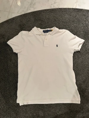 Vit piké från Polo Ralph Lauren - Tjena, säljer min vita pikétröja från Polo Ralph Lauren i slim fit-modell. Klassisk krage med två knappar och den ikoniska broderade loggan på bröstet. Tillverkad i mjuk bomull som är skön mot huden. Perfekt för dig som gillar stilrena och tidlösa plagg.