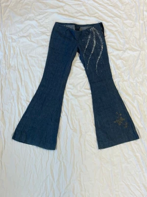 Blå bootcut jeans med glitterdetaljer - Snygga blå bootcut jeans med glittrande paljettdetaljer längs ena benet och ett guldfärgat blommigt tryck vid benslutet. Jeansen har låg midja och är tillverkade i klassiskt jeansmaterial. Perfekt för dig som vill sticka ut med coola detaljer och unik stil.