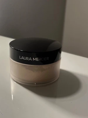 Laura Mercier Translucent Loose Powder - Translucent Loose Setting Powder från Laura Mercier i en rund plastburk med svart lock. Pudret är ljust och transparent, perfekt för att fixera smink och ge en matt finish. Förpackningen har sifter för enkel dosering och innehåller 29g.