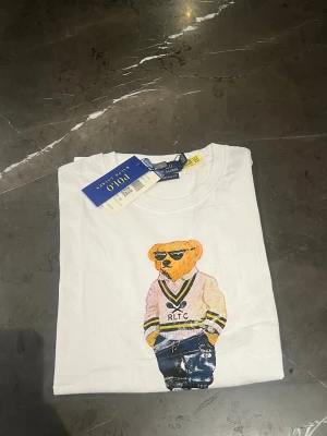 Vit Polo Bear t-shirt från Ralph Lauren - Säljer en vit t-shirt från Ralph Lauren med Polo Bear-tryck på framsidan. Björnen har solglasögon, stickad tröja och jeans, vilket ger en chill vibe. T-shirten har rund hals och korta ärmar. Materialet är mjuk bomull och passformen är normal.