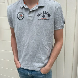 Grå piké från Sail Racing - Säljer en grå pikétröja från Sail Racing med korta ärmar och klassisk krage. Tröjan har tryck och broderade detaljer på bröstet, samt en patch på ärmen. Materialet är mjuk bomull och insidan av kragen har orange detaljer. Perfekt till jeans eller shorts.