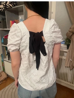 Vit blus med svart rosett och blommig struktur - Säljer en vit blus med korta puffärmar och fyrkantig halsringning. Blusen har ett blommigt, broderat mönster och en stor svart rosett i ryggen som knyts. Materialet känns som bomull eller polyester och toppen har en feminin och söt vibe. Från shein men riktig bra material💕