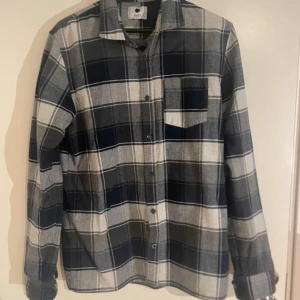 Rutig skjorta från NN07 Pedro - Säljer en snygg rutig skjorta från NN07, modell Pedro. Skjortan är i mörkblått, grått och vitt med klassisk krage, bröstficka och knappar framtill. Tillverkad i 100% bomull, perfekt för dig som gillar stilrena och bekväma plagg.