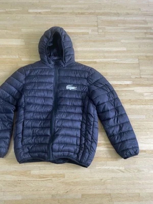 Svart pufferjacka från Lacoste - Säljer en svart pufferjacka från Lacoste med huva och dragkedja framtill. Jackan har det klassiska Lacoste-logotypen på bröstet och är quiltad för extra värme. Perfekt för kyliga dagar och har en clean, sportig look.