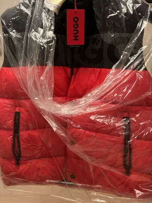 Röd och svart dunväst från Hugo - Säljer en riktigt snygg dunväst från Hugo i rött och svart. Västen har två praktiska fickor med svarta dragkedjor och ett coolt blockmönster. Perfekt att slänga över en hoodie när det är kyligt ute. Märket syns tydligt framtill.