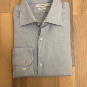 Ljusblå slim fit skjorta J.Lindeberg - Snygg ljusblå skjorta från J.Lindeberg i slim fit-modell. Tillverkad i ett tunt och mjukt bomullstyg som känns fräscht och behagligt mot huden. Skjortan har klassisk krage, vita knappar och långa ärmar med manschett. En stilren modell som ger en clean och modern look.  Mått: 	•	Armhåla till armhåla: 46 cm 	•	Axel till axel: 52 cm 	•	Topp till botten: 83 cm 	•	Axel till ärmslut (cuff): 66 cm