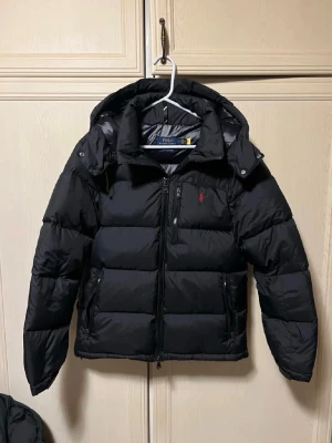 Svart pufferjacka från Polo Ralph Lauren - Svart pufferjacka från Polo Ralph Lauren med huva och dragkedja framtill. Jackan har broderad röd logga på bröstet och två sidofickor med dragkedja. Materialet är polyester och jackan är perfekt för kalla dagar.