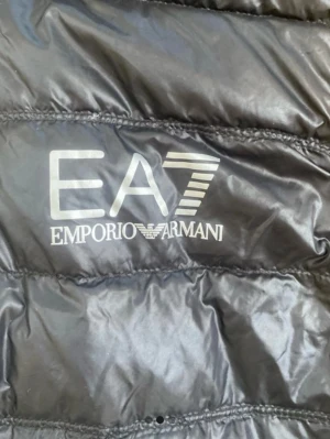 Svart pufferjacka från Emporio Armani - Svart pufferjacka från Emporio Armani EA7 med glansig finish och tydlig logga på bröstet. Jackan har dragkedja framtill med EA7-detalj på zip-pullern. Perfekt för dig som vill ha en snygg och modern look.