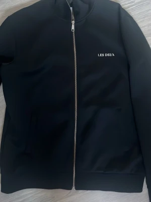 Svart zip-kofta från Les Deux - Svart kofta från Les Deux med hel dragkedja och diskret logga på bröstet. Tröjan har lång ärm, ståkrage och två fickor framtill. Materialet känns mjukt och har en clean look som funkar till många stilar. Perfekt för dig som gillar minimalistiskt och stilrent. Priset är inte hugget i sten så det är bara att höra av sig vid frågor 