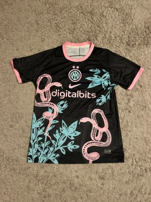 Inter special edition tröja - Bilderna visar en svart Inter Milan x Nike fotbollströja med rosa och turkosa detaljer, inklusive ormmönster och blommor. Tröjan har korta ärmar och Digitalbits-logga på bröstet. 