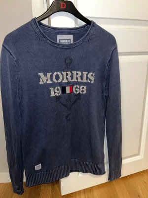 Blå stickad tröja Morris 1968 - Säljer en blå stickad tröja från Morris med broderad text 'Morris 1968' och ett ankare på bröstet. Tröjan har rund hals, ribbade muddar och lång ärm. Perfekt för dig som gillar klassisk stil med en twist.