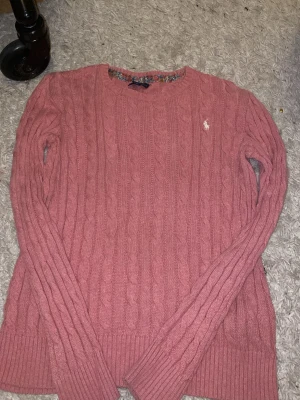 Rosa stickad tröja från Polo Ralph Lauren - Säljer en rosa kabelstickad tröja från Polo Ralph Lauren i storlek XL. Tröjan har långärmad design, rund hals och den klassiska broderade loggan i vitt på bröstet. Materialet är mjuk bomull och passformen är normal. Perfekt för dig som gillar stilrena och bekväma plagg.