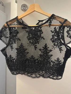 Svart spetsblus från NA-KD - Säljer en svart, transparent blus från NA-KD i storlek 36. Blusen har korta ärmar och är dekorerad med broderade blommor i svart spets över hela plagget. Den är croppad och har en rund halsringning med fina detaljer längs kanten.