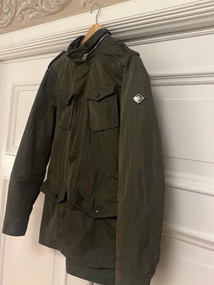 J.lindberg fieldjacket - Snygg mörkgrön parkajacka från J.Lindeberg med hög krage, dragkedja och knappar framtill. Jackan har flera fickor med lock och coolt emblem på ärmen. Materialet är polyester och jackan har långärmad design, perfekt för höst och vinter.