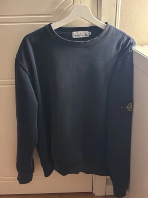 Svart sweatshirt från Stone Island - Säljer en svart långärmad sweatshirt från Stone Island med klassisk rund hals och ikonisk patch på vänster ärm. Tröjan har en loose passform och är tillverkad i mjuk bomull, perfekt för chill dagar. Enkelt och stilrent plagg som lyfter din streetwear-look.