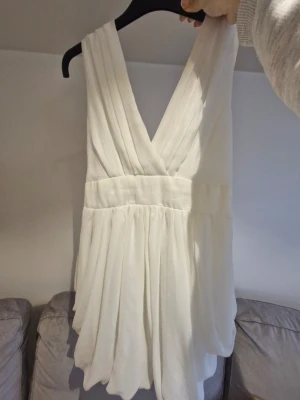 Vit omlottklänning med v-ringning - Säljer en vit kortklänning med omlott och djup v-ringning både fram och bak. Klänningen har breda axelband och ett smockat midjeparti baktill som ger en snygg passform. Materialet är lätt och luftigt, perfekt för varmare dagar.