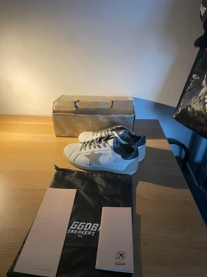 Golden goose Superstar sneakers - Säljer ett par GGDB Superstar sneakers i vitt skinn med silverfärgad stjärna på sidan och mörkblå häl. Skorna har grå snörning, rund tå och platt sula. Medföljer originalpåse och kartong. Perfekt för dig som gillar streetstyle och vill ha något unikt.