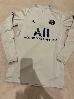 Vit Nike air Jordan psg sport tröja storlek S - Bilderna visar en långärmad träningströja från Paris Saint-Germain x Jordan, inte skor. Tröjan är ljusgrå med blå och röda detaljer, halv dragkedja och loggor på bröstet och ärmarna. Materialet är polyester och elastan.