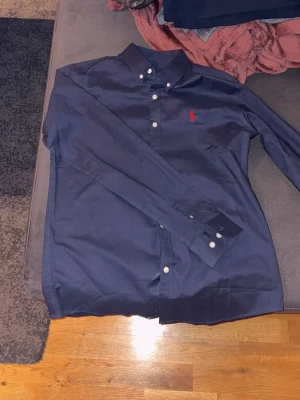 Marinblå skjorta från Ralph Lauren - Säljer en marinblå skjorta från Ralph Lauren i klassisk passform. Skjortan har långa ärmar, knappar framtill och en röd broderad logga på bröstet. Materialet är mjuk bomull och kragen har knappar för extra stilpoäng.