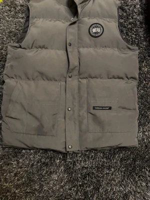 Grå dunväst från Canada Goose - Säljer en grå dunväst från Canada Goose med klassisk logga på bröstet och två stora fickor framtill. Västen har tryckknappar och är tillverkad i slitstark polyester med dunfyllning. Insidan har meshficka och etikett med QR-kod. Perfekt för kyliga dagar.