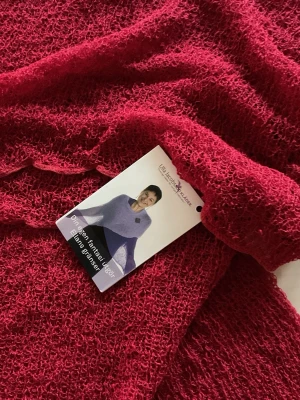 Röd stickad cape från Ulla Jacobsson - Säljer en röd stickad cape från Ulla Jacobsson som kan stylas på flera olika sätt. Capen har en luftig och grovstickad struktur med vågig kant och kan bäras både som axelvärmare eller poncho. Perfekt för att ge din outfit en färgklick och extra värme.