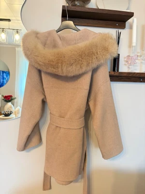 Beige kappa med pälskant och bälte - Snygg beige kappa med fluffig pälskant på huvan och knytbälte i midjan. Jackan har en avslappnad passform och långa ärmar, perfekt för dig som vill ha en mysig och trendig look. Materialet känns mjukt och värmande, och huvan ger extra stilpoäng.