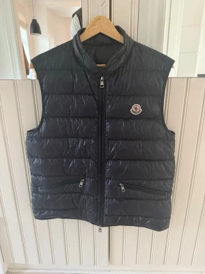 Blå Moncler Gui väst  - Säljer denna mörkblå moncler gui västen i storlek 4. Den är mycket sparsamt använd (nyskick) och det finns kvitto samt tags kvar. Nypris idag är ca 9000 kr. Hör av dig om du vill ha fler bilder eller har frågor! 