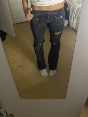 Svarta slitna bootcut jeans från gina - Säljer ett par svarta bootcut jeans med slitningar över knäna som jag har klippt upp själv och fickdetaljer framtill. Lågmidjade och i klassisk jeanskvalitet med dragkedja och knapp. 