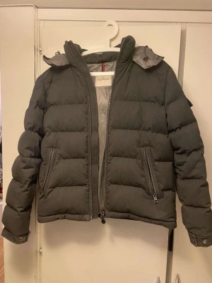 Mörkgrå pufferjacka från Moncler - Säljer en mörkgrå pufferjacka från Moncler med huva och två sidofickor med dragkedja. Jackan har ett stilrent, quiltat mönster och Moncler-logga på ärmen. Perfekt för kalla dagar och har en snygg, modern look.