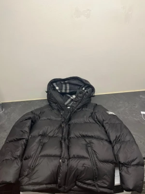 Svart pufferjacka  - Säljer en svart pufferjacka  med klassisk logotyp på ärmen och rutigt foder i huvan. Jackan har dragkedja och knappar framtill, två sidofickor med dragkedja och en skön, vadderad känsla. Perfekt för kalla dagar och riktigt snygg stil.