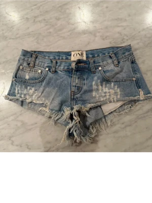 Blå denimshorts från ONE Trash Whores - Säljer ett par blå denimshorts från ONE Trash Whores med råa, fransiga kanter och slitna detaljer framtill och baktill. Klassisk femficksmodell med knapp och dragkedja. Perfekt för dig som gillar en edgy och avslappnad stil.