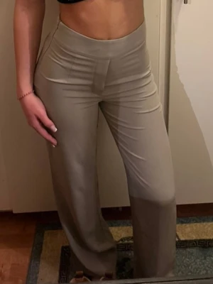 Beiga wide kostymbyxor med hög midja - Snygga beiga kostymbyxor med hög midja och wide fit. Byxorna har en slät front utan synliga fickor och är tillverkade i ett mjukt, lätt material som ger en clean och stilren look. Perfekta att matcha med crop tops eller skjortor för en trendig vibe.