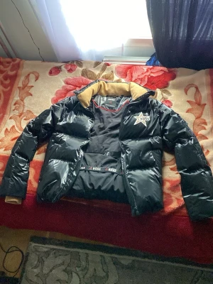 Svart pufferjacka från BOSS - Svart glansig helt ny äkta pufferjacka från BOSS med stor beige BOSS-logga på ryggen och stjärndetalj på bröstet. Jackan har beige muddar och krage, dragkedja framtill och BOSS-band på insidan. Perfekt för dig som vill sticka ut med en fet jacka.