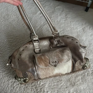 Italiensk guld handväska med cowprint detalj - Unik handväska i guldigt skinn med präglade blommönster och detaljer i päls på framsidan. Väskan har två handtag, dekorativa nitar och en ficka med paisleymönstrat foder i grönt och svart. Dragkedja upptill och flera fack inuti.