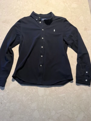 Mörkblå skjorta från Polo Ralph Lauren - Snygg mörkblå skjorta från Polo Ralph Lauren med klassisk krage och vita knappar framtill. Skjortan har lång ärm och den ikoniska broderade loggan på bröstet. Tillverkad i mjuk bomull som är skön mot huden. Perfekt för dig som gillar stilrena och tidlösa plagg. Pris 379kr storlek xs!