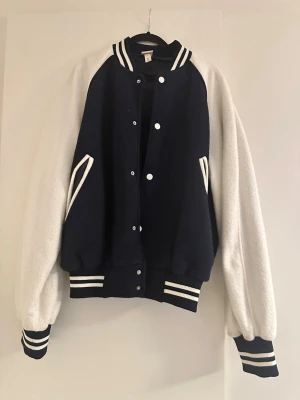 Blå och vit bomberjacka från H&M - Säljer en snygg bomberjacka i mörkblått med vita ärmar och vita detaljer. Jackan har ribbade muddar och krage med ränder, tryckknappar framtill och två fickor. Materialet är en mix av polyester och bomull, perfekt för dig som vill ha en sportig och cool look.