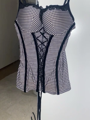 Svartvit rutig korsett med snörning - Säljer en snygg korsettopp i svartvitt rutigt mönster med svarta detaljer och spetskant upptill. Toppen har snörning framtill och justerbara axelband. Perfekt för dig som vill ha en statement piece med cool vintagevibe.