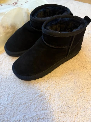Svarta boots i fårskinn, storlek 38 - Mysiga svarta boots med rund tå och platt sula, fodrade med mjukt fårskinn på insidan. Utsidan är i mocka och sulan är rejäl med mönster för extra grepp. Perfekta för kalla dagar och riktigt sköna att dra på sig när det är vinter.